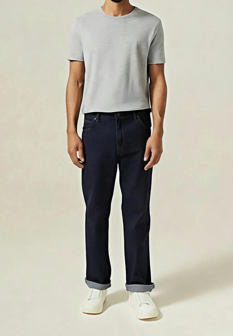 Homme portant un t-shirt gris clair à manches courtes, un jean foncé retroussé et des chaussures blanches, debout devant un fond uni.