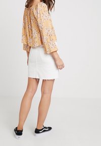 Kvinde iført en gul blomsterlangærmet blouse, hvid denim mini nederdel med rå kant og sorte lavtop sneakers, stående med ryggen til.