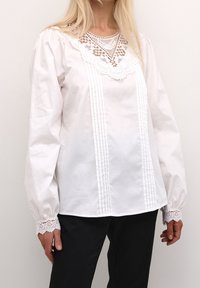 Bílá blůza s dlouhými nakládanými rukávy, krajkovými detaily na výstřihu a manžetách, s nařaseným předním dílem a volnějším střihem.