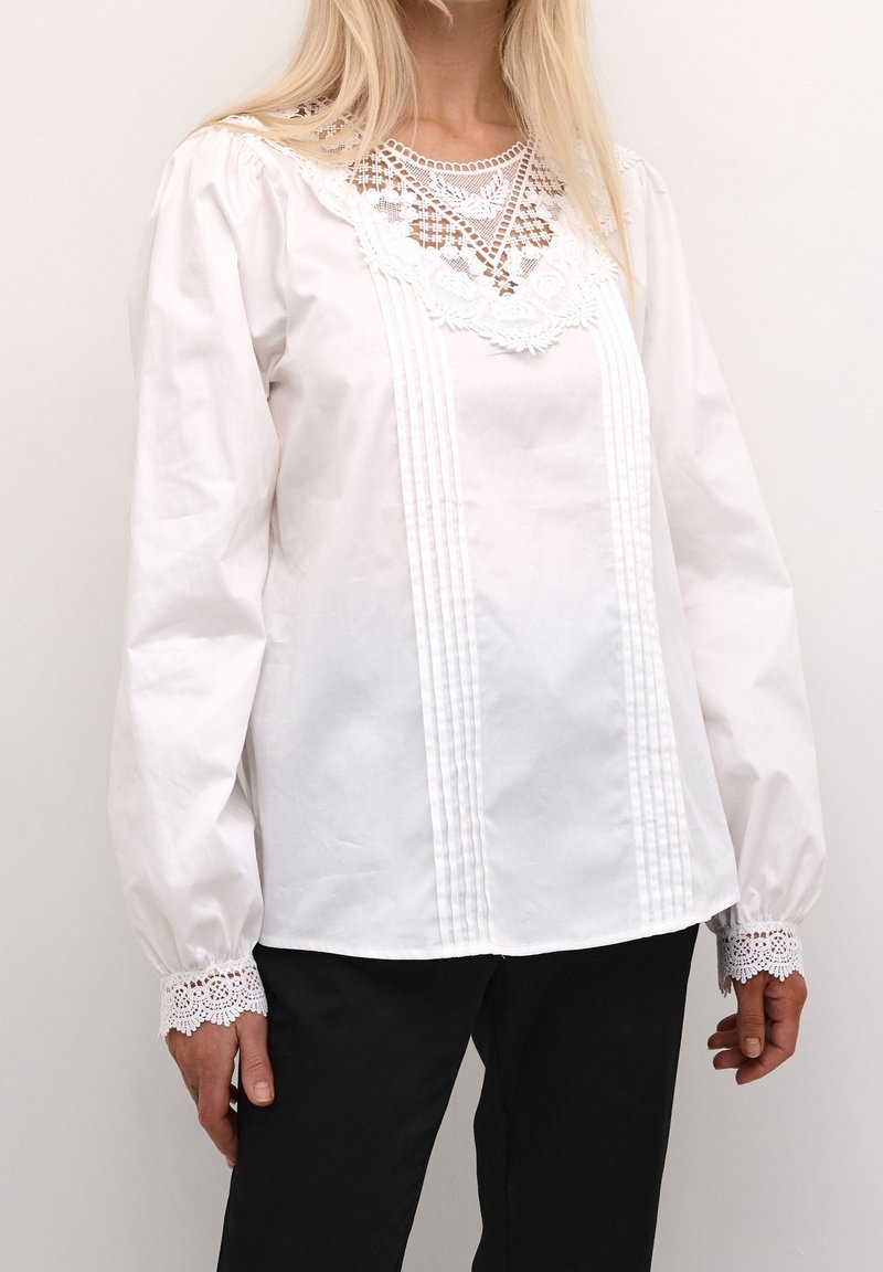Bílá blůza s dlouhými nakládanými rukávy, krajkovými detaily na výstřihu a manžetách, s nařaseným předním dílem a volnějším střihem.