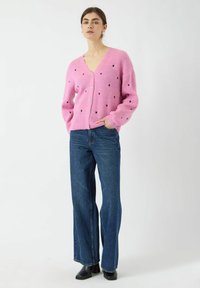 Rosa gestrickte Strickjacke mit Herzmuster-Ausschnitten und vorderen Knöpfen, kombiniert mit blauen weiten Jeans und schwarzen Ankle Boots.
