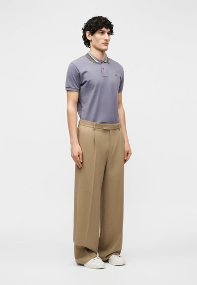 Jeune homme debout, portant un polo violet avec col rayé, un pantalon beige à jambes larges, et des baskets blanches sur un fond uni.