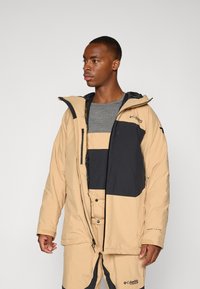 Beige vinterjakke med svart og kremfarget fargebånddesign, laget av vanntett materiale med hette og glidelås foran.