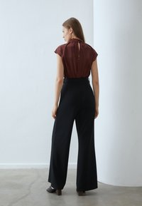 Bruine blouse met korte mouwen, hoge hals en een sleutelgat aan de achterkant, gecombineerd met zwarte hoge taille wijde broek, gezien vanuit de achterkant.