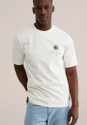 Mann trägt ein weißes T-Shirt mit einem grünen Palmenmotiv und dem Schriftzug "The Leisure Court Club", kombiniert mit hellblauen Jeans vor einem schlichten Hintergrund.