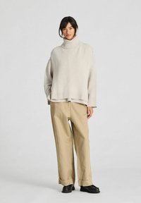 Mujer de pie vistiendo un suéter de cuello alto beige, pantalones caqui holgados con puños y zapatos negros, sobre un fondo claro y liso.