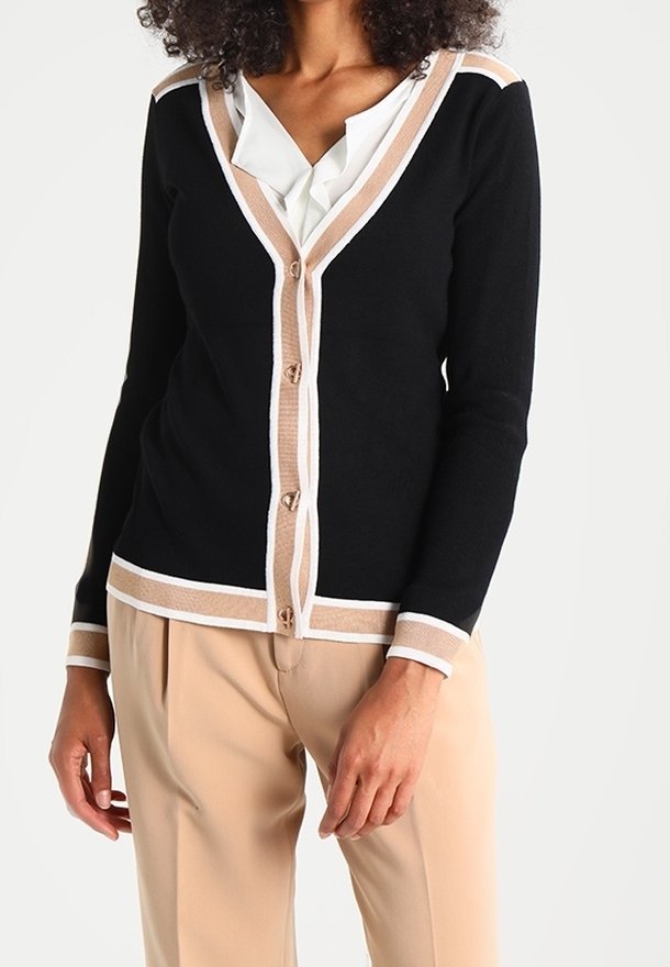 Černý cardigan s bílými a béžovými pruhy, má V-výstřih a zapínání na knoflíky, vyrobený z měkkého pleteného materiálu, kombinovaný s světlými béžovými kalhotami.