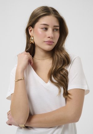 Jeune femme aux longs cheveux bruns ondulés porte un t-shirt blanc à col en V, de grandes boucles d'oreilles dorées en forme de créoles, un bracelet en or et un collier en or délicat, regardant sur le côté.