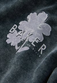 Tumša auduma ar gaiši pelēku ziedu rakstu un uzrakstu "BAKER" izcili redzamu. Tekstūra šķiet mīksta un viegli raiba.