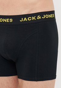 Nahaufnahme einer Person, die schwarze Boxershorts mit einem schwarzen Bund trägt, auf dem der gelbe Schriftzug "JACK & JONES" zu sehen ist.