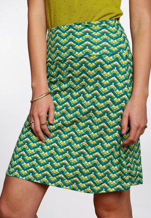 Gros plan sur une femme portant une jupe à motifs géométriques verts et jaunes, associée à un haut vert texturé et des bijoux dorés à ses mains.
