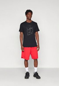 Camiseta preta de algodão com um grande gráfico cinzento e logótipo da Nike, combinada com calções vermelhos brilhantes e ténis pretos. Meias brancas visíveis.