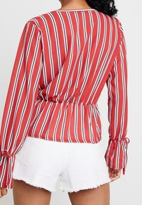 Rot-weiß gestreifte Bluse mit langen Ärmeln, geraffter Taille und Rüschendetails an den Ärmelabschlüssen, kombiniert mit weißen Fransen-Shorts.