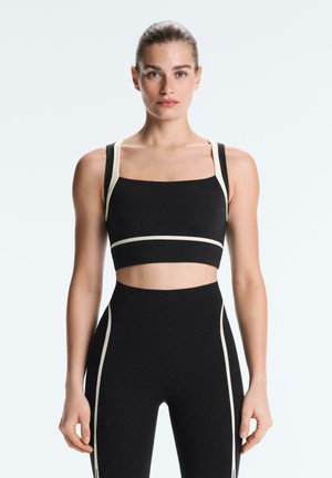 Nő fekete és bézs sportmelltartót és magas derekú leggingset visel, szemben áll egy világos háttér előtt.
