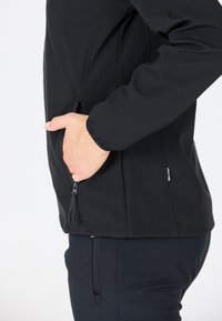 Veste softshell noire avec poches zippées, design ajusté et tissu texturé. Couture détaillée et accent de logo sur le côté.