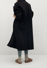 Abrigo largo negro con cuello, ajuste relajado, tejido texturizado, combinado con leggins azul claro y zapatillas beige con calcetines de punto.