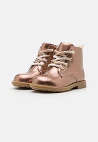 Botas metálicas de color rosa dorado, con cordones, de tamaño infantil, con suelas beis sobre fondo blanco, inclinadas ligeramente a la derecha.