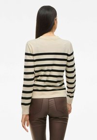 Femme avec de longs cheveux bruns portant un pull beige à rayures noires horizontales et un pantalon moulant brun, tournée vers le dos sur un fond blanc.