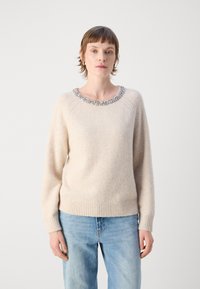 Pull en tricot beige avec une finition texturée et un col orné de perles. Présente des poignets et un ourlet côtelés, porté avec un jean bleu clair.