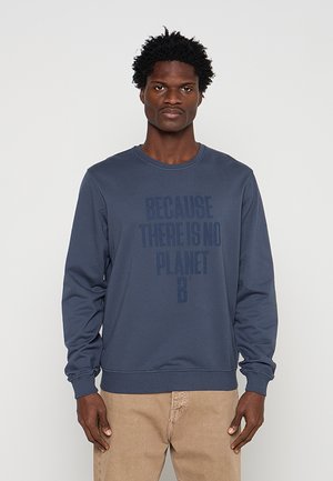 Hombre con sudadera azul con el texto «Because there is no Planet B», de pie contra un fondo claro y liso.
