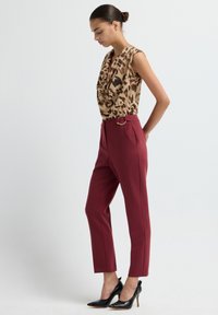 Woman standing in leopard-print sleeveless top, high-waist burgundy trousers with belt detail, and black pointed high heels, against plain background.