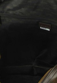 Interno di una borsa in tessuto nero con una tasca con cerniera, una targhetta con il logo Guess in pelle marrone e un'etichetta GuessEco.