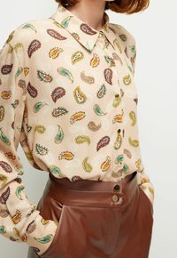 Blusa de manga larga en tejido beige con coloridos patrones de paisley. Brillo sutil, cuello clásico y detalle de botonera. Combinada con pantalones marrones.