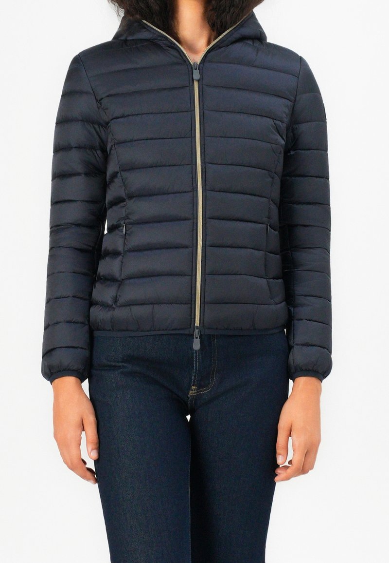 Femme portant une veste matelassée noire zippée avec capuche et un jean bleu foncé, sur un fond blanc uni.