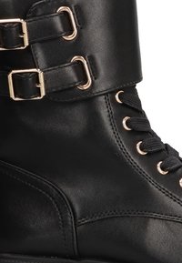 Botín de cuero negro con hebillas en tono dorado, cierre con cordones en la parte delantera y superficie texturizada. Presenta una puntera redondeada y detalles de costura.
