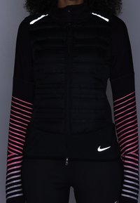 Černá péřová vesta se zipem vpředu, která má bílé reflexní detaily. Dlouhé rukávy v černé barvě s růžovým pruhovaným vzorem. Logo Nike v bílé barvě.