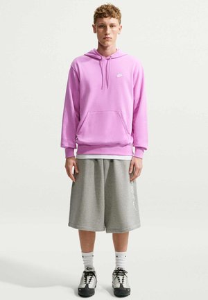 Młody mężczyzna w różowej bluzie z kapturem Nike, szarych szerokich spodenkach, białych skarpetkach i czarno-białych trampkach stoi na jednolitym tle.
