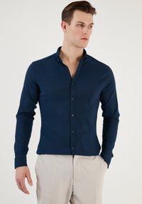 Cămașă albastră închis cu mâneci lungi din țesătură texturată, având un guler cu nasturi, cu un design ajustat și nasturi vizibili pe față.