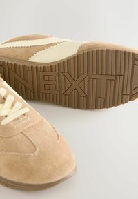 Zīda zamša bēšā krāsā sporta apavi ar krēmkrāsas akcentiem, apaļu purngalu, auklu aizdari un teksturētu brūnu gumijas zoli ar reljefā iespiestu tekstu.