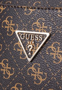 Materiale nero testurizzato con un motivo a monogramma dorato. Placchetta centrale triangolare in metallo con "GUESS USA" e logo con punto interrogativo.