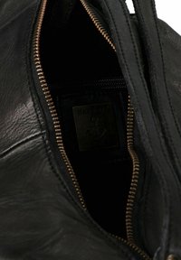 Innenansicht einer schwarzen Lederhandtasche, die einen bronzefarbenen Reißverschluss und eine Materialstruktur aufweist, mit einem Etikett, das mit einem Ankersymbol geprägt ist.