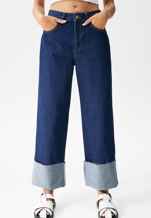 Un jean large bleu foncé présente un ourlet effilé avec un revers en denim plus clair. Les détails comprennent des poches avant et une fermeture à bouton. Porté avec des sandales blanches.
