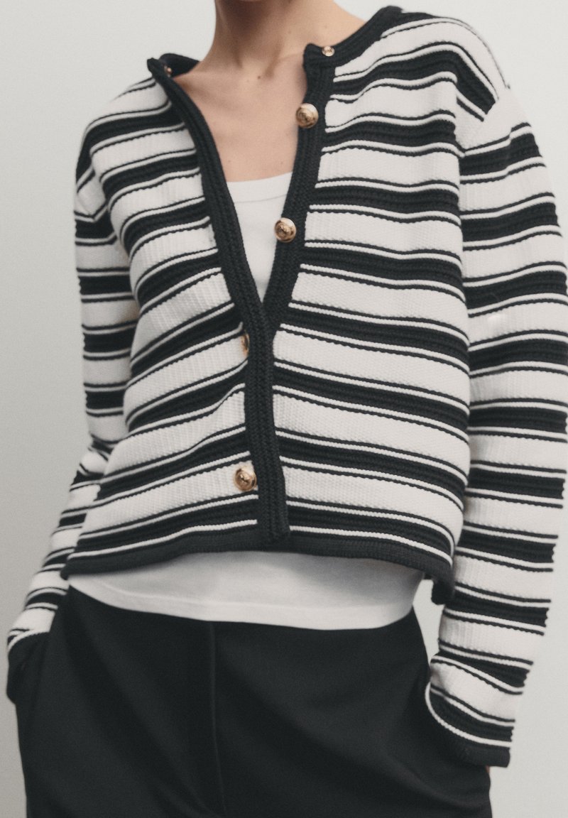 Personne portant un cardigan rayé horizontal noir et blanc avec des boutons dorés sur un haut blanc, associé à un pantalon noir, mains dans les poches.