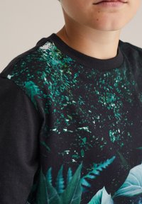 Mustat hupparit, joissa on edessä värikäs lehtikuvio vihreissä ja sinisissä sävyissä, mustilla teksturoiduilla kankailla hihoissa ja kauluksessa.