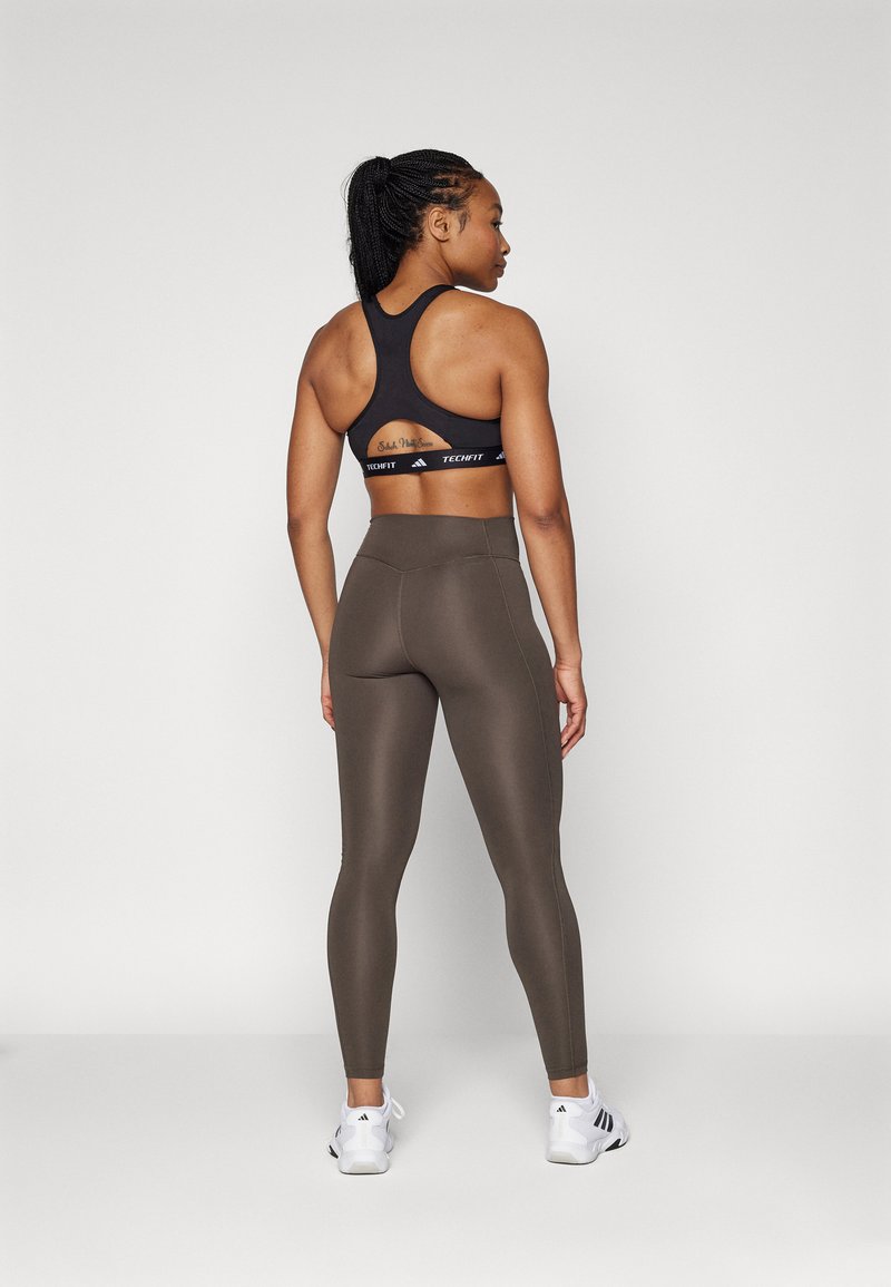 Soutien-gorge de sport noir avec un design dos nageur, associé à un legging olive à taille haute et lisse, et des chaussures de sport blanches.