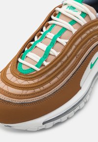 Gros plan d'une basket marron et beige avec des lacets blancs, des œillets verts, des coutures vertes et une semelle blanche avec un coussin d'air visible.