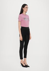 T-shirt rosa a maniche corte con stampa floreale e testo "COUTURE", abbinato a leggings neri aderenti e tacchi alti neri, in posa in piedi.