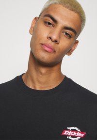 Camiseta de algodón negra con cuello redondo y un logo rojo y blanco en el lado izquierdo. Textura suave, diseño casual.