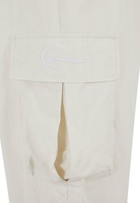 Beige stoff med en lomme som har hvit brodert "Kane." Designet inkluderer en plissering under lommen.
