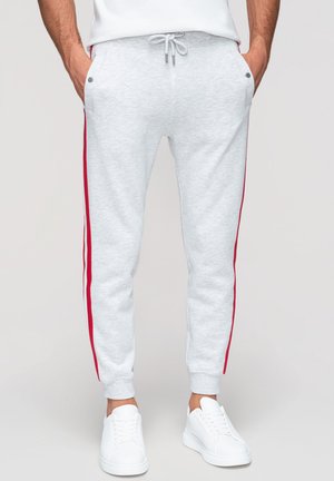Homme portant un jogging gris clair avec des bandes rouges sur les côtés, des baskets blanches, les mains dans les poches, debout devant un fond uni.