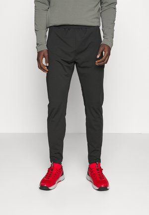 Mann trägt schwarze, schmal zulaufende Hose, graues Langarmshirt und leuchtend rote Sneakers und steht vor weißem Hintergrund.