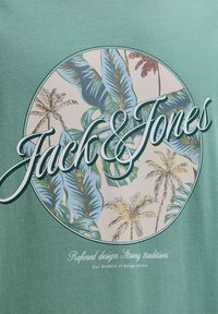 Tela verde con un diseño circular de hojas de palma tropical y el texto estilizado "Jack & Jones" en el centro.
