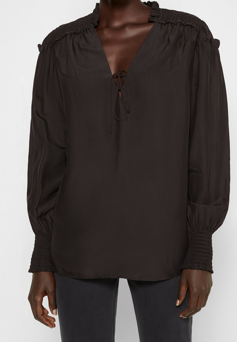 Blouse noire avec un profond décolleté en V et un lien ajustable. Caractérisée par des détails froncés sur les épaules et les poignets, des manches longues, et une coupe décontractée.