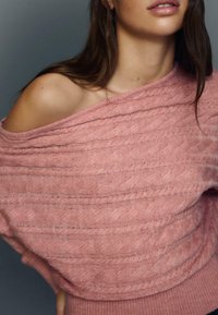 Lyserød strikket sweater med løs, skulderfri pasform. Har tekstureret mønster og ribbet halsudskæring, som fremhæver blødhed og varme.