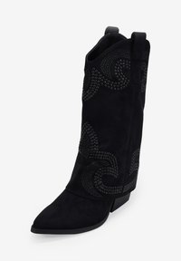 Botas cowboy de ante negro con punta afilada, tacón corto en bloque y un diseño intrincado de tachuelas en el caña. Detalles del patrón texturizado visibles.