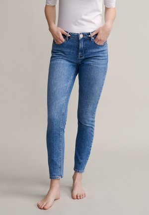 Femme portant un jean skinny bleu clair et un haut blanc, debout pieds nus les mains dans les poches, sur un fond uni.